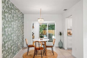 A dining area with a round table, four chairs, a hanging light fixture, and a view of a garden through a glass door.