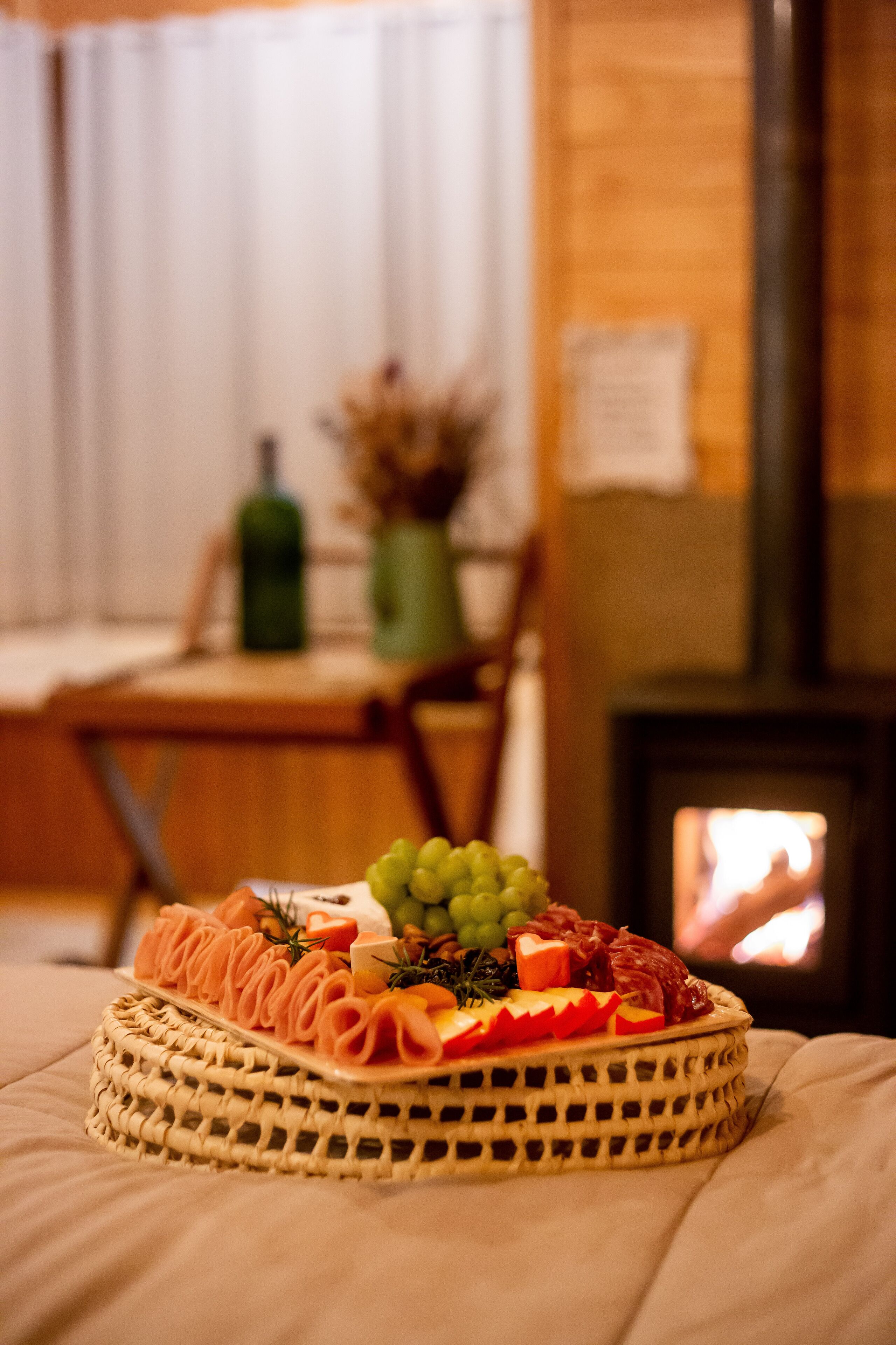 A round fruit and cheese platter on a bed with a wooden headboard and a fireplace in the background.