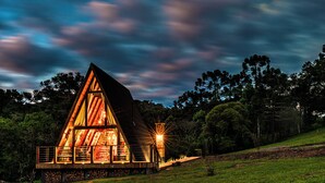 A triangular wooden cabin with a glass facade, surrounded by trees and a grassy area. - Urutau Pousada (Urubici)