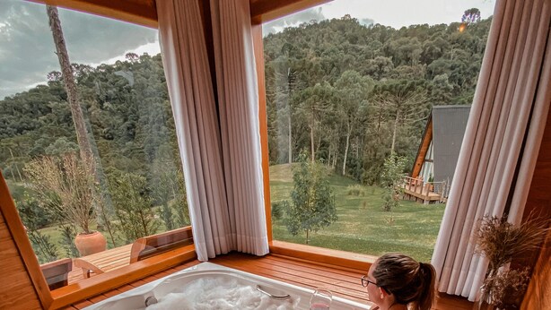 A wooden cabin with a large bathtub filled with bubbles, a wooden deck, and a view of a forested hill. - Urutau Pousada (Urubici)
