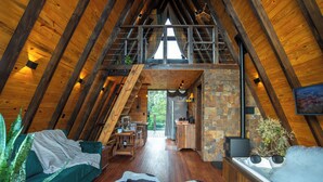 A cozy, rustic interior with a sloped ceiling, wooden beams, a stone fireplace, a green sofa, a cowhide rug, and a hot tub. - Urutau Pousada (Urubici)