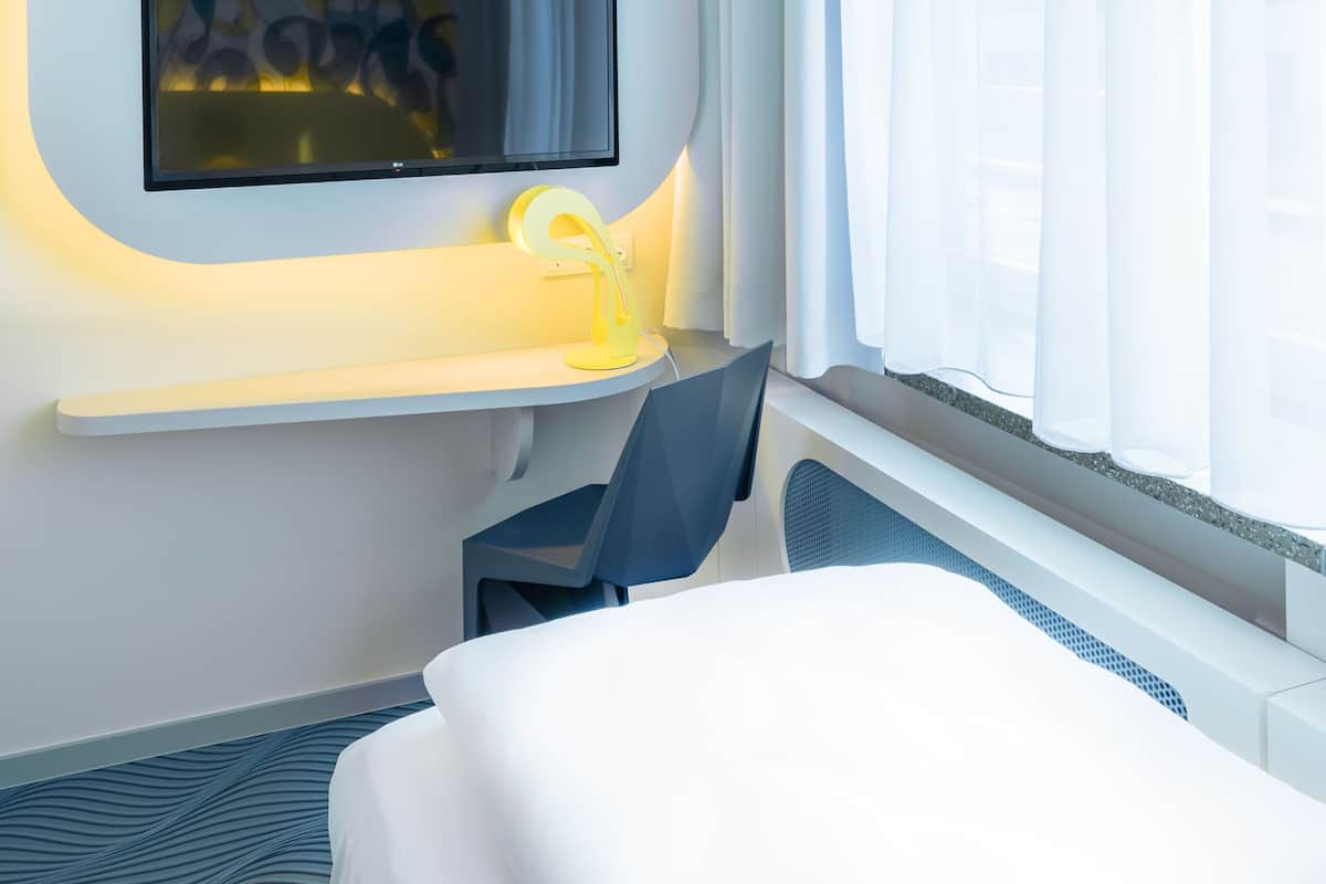 Une chambre d’hôtel comprenant un lit, un bureau avec une chaise, un téléviseur à écran plat fixé au mur et une fenêtre avec des rideaux.