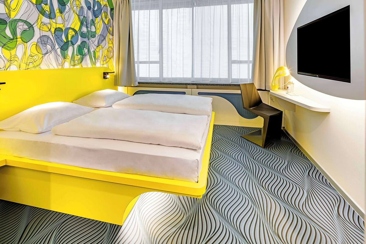 Une chambre d’hôtel avec un lit, un bureau, une télévision et une fenêtre avec des rideaux.