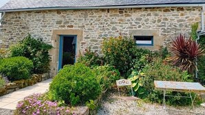 A stone house with a tiled roof, a small porch, and a garden with various plants and flowers.
