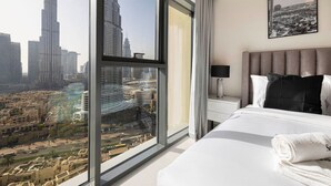 A hotel room with a large window offering a cityscape view, a neatly made bed, a bedside table with a lamp, and a framed picture on the wall. - New 2B | Burj Khalifa | Dubai Mall | 2 Pools | Gym (Dubai)
