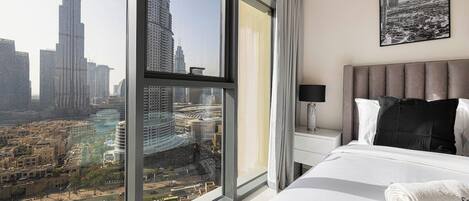 Habitación de hotel con un ventanal amplio que ofrece vistas a un paisaje urbano, una cama, una mesita de noche y un cuadro en la pared.
