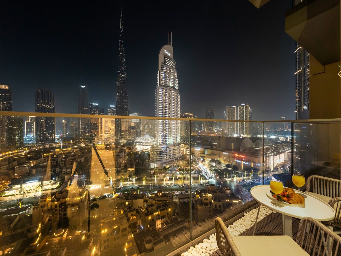 A high-rise hotel room with a panoramic city view, a dining table with a fruit bowl, and a glass railing.