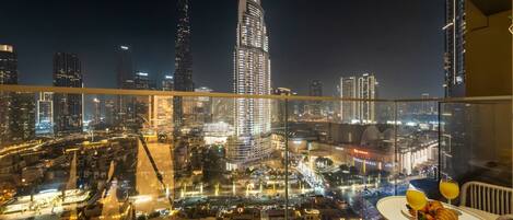 A high-rise hotel room with a panoramic city view, a dining table with a fruit bowl, and a glass railing.