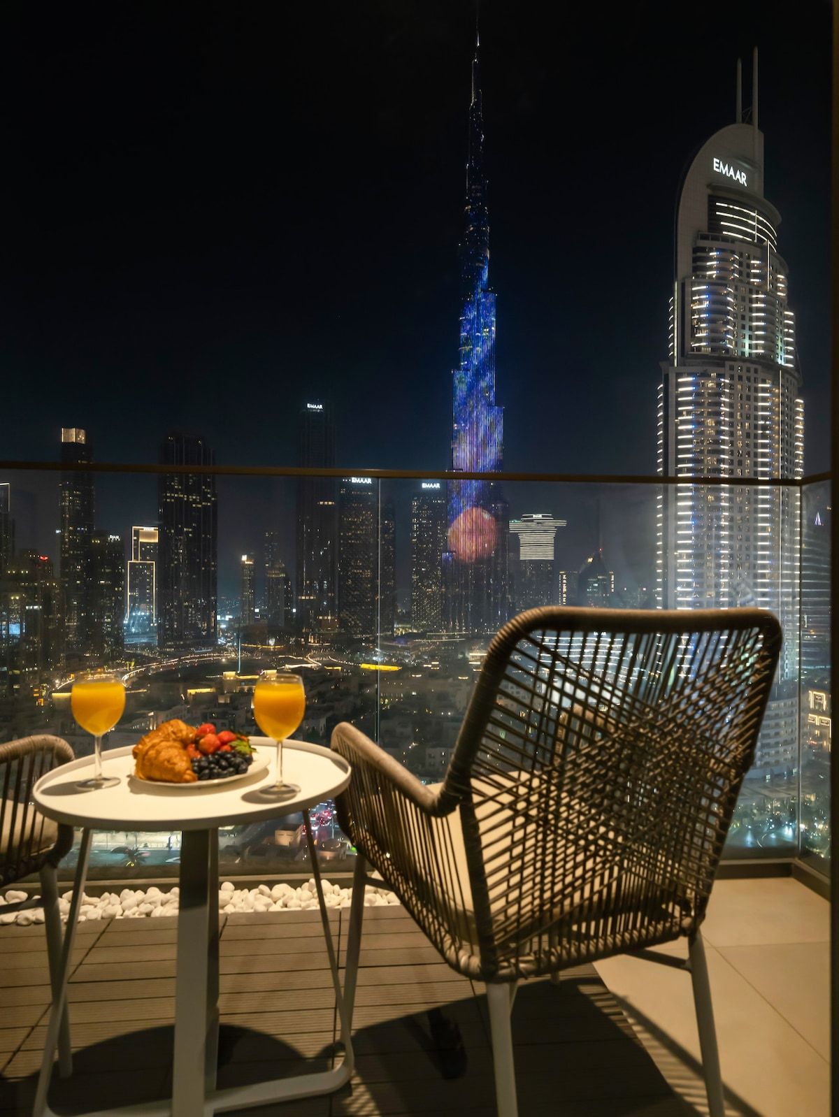 Un balcon avec une table et des chaises, d’où l’on peut admirer un panorama sur la ville, avec ses bâtiments illuminés, dont une tour imposante.