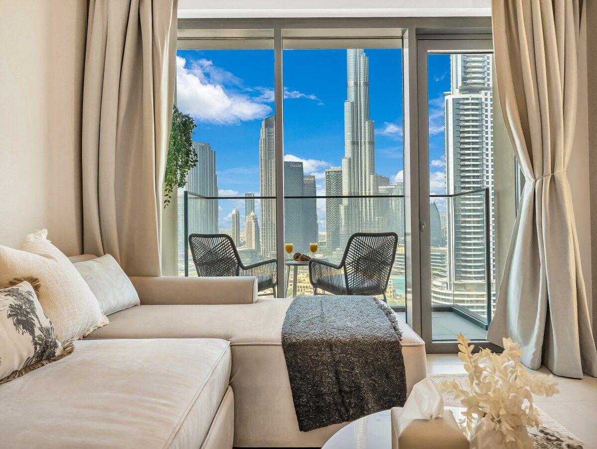 A modern hotel room with a large balcony, a sofa, two chairs, and a table with a city view.