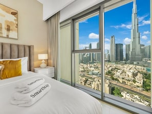 A hotel room with a large window offering a city view, a bed with white linens, a nightstand with a lamp, and a painting on the wall. - New 2B | Burj Khalifa | Dubai Mall | 2 Pools | Gym (Dubai)