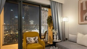 Un salon moderne doté d’une grande fenêtre donnant sur une vue nocturne de la ville, avec un fauteuil jaune, un canapé gris et une petite table d’appoint surmontée d’une lampe.