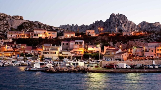 A coastal town with buildings on a hillside, a marina with boats, and rocky cliffs in the background.