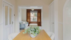 A dining area with a wooden table, white chairs, a potted plant, and a view through a window with wooden shutters. - Prime Location 2 Bed East London (London)