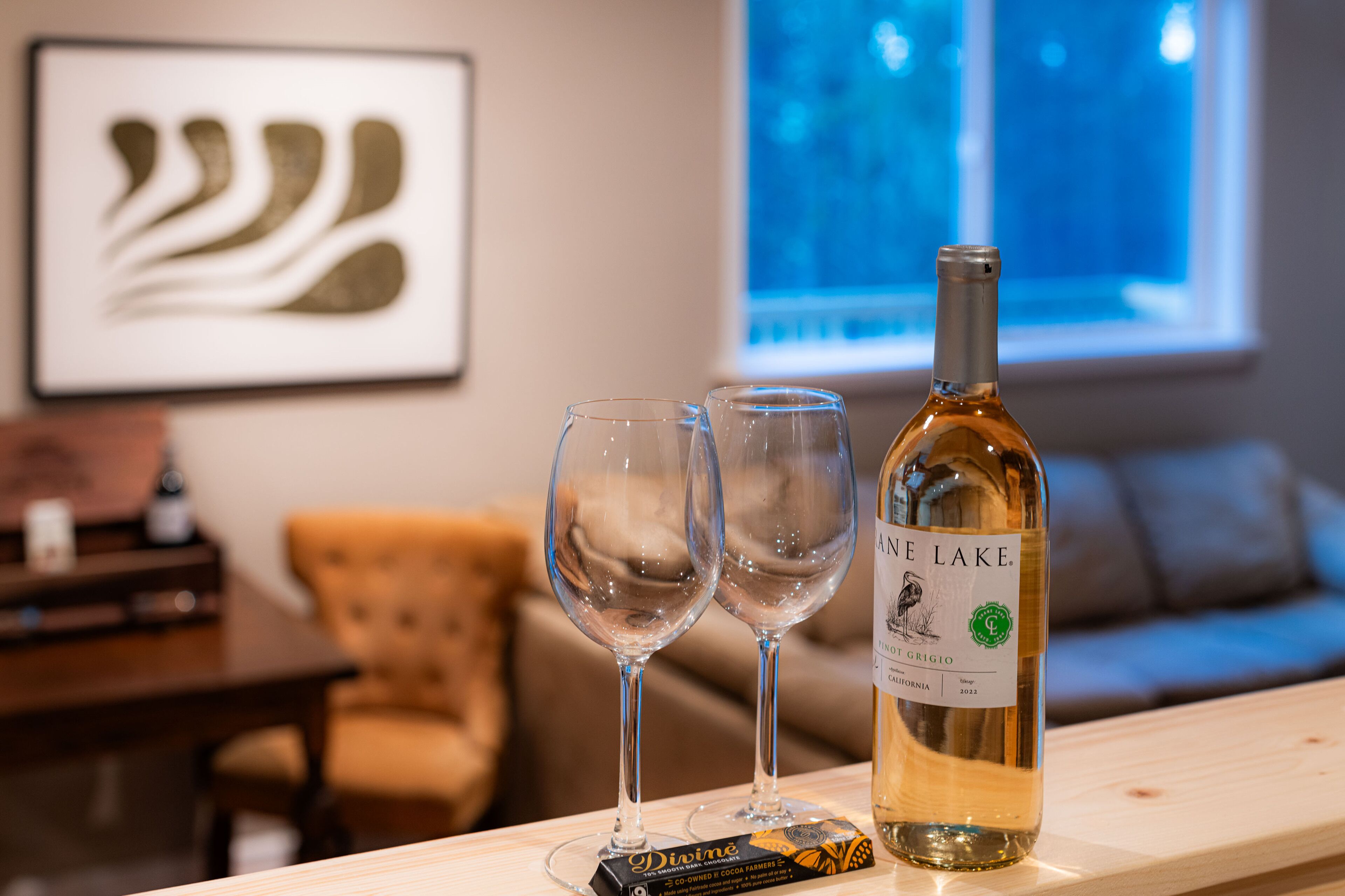 A bottle of wine and two glasses on a wooden table in a room with a couch and a framed picture on the wall.