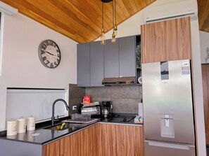 A modern kitchen with wooden cabinets, a built-in oven, a coffee maker, and a clock on the wall. - Luxury Glass Cabin- 270° Ocean & Forest Views + Hot Tub (Provincia de Puntarenas)