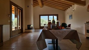 A spacious room with a high ceiling and exposed wooden beams, a large window with a view of the outdoors, a table set with food and drinks, and a radiator on the floor.