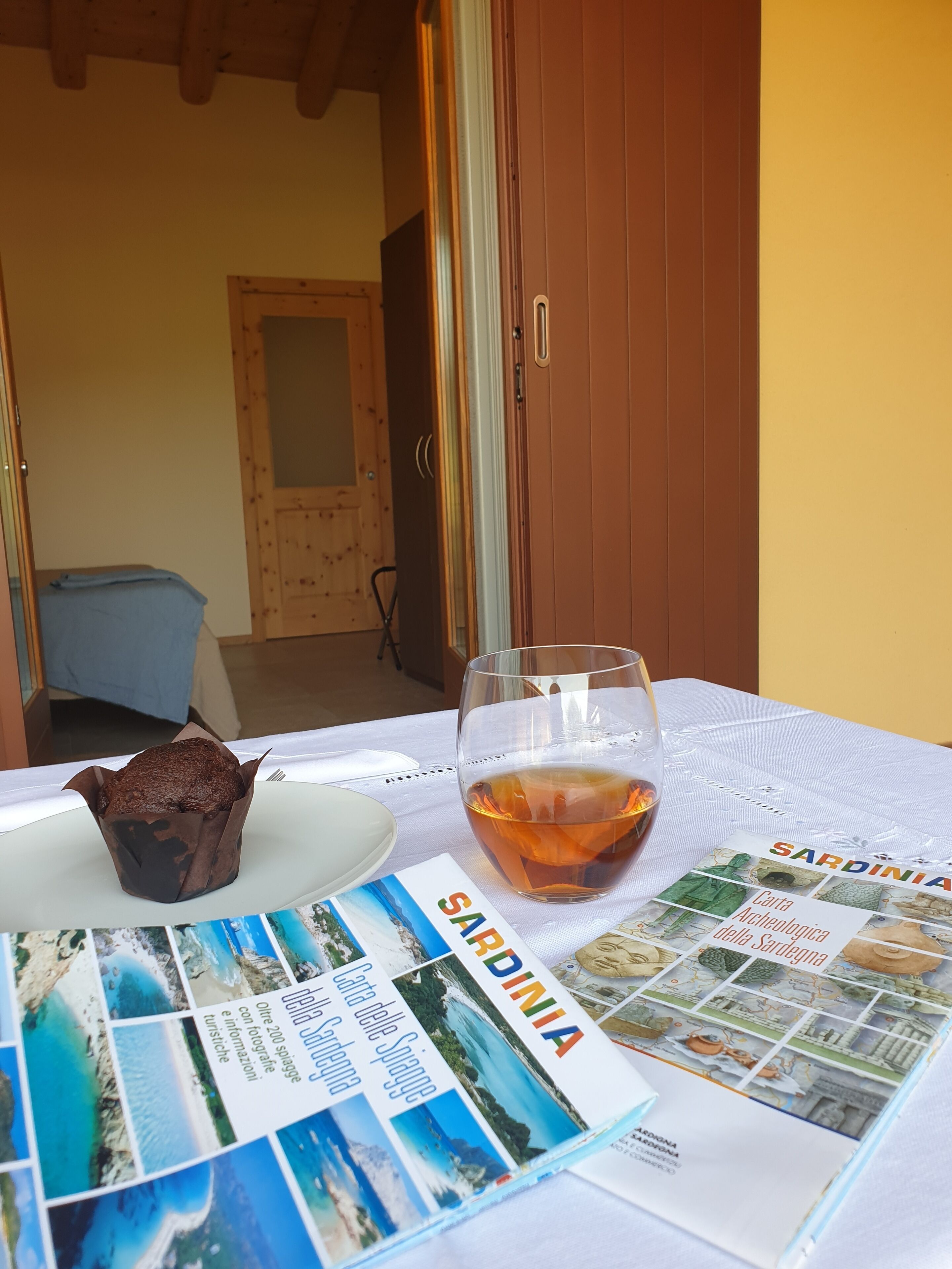 A table with a glass of amber liquid, a muffin, and a travel brochure titled "SARDINIA".