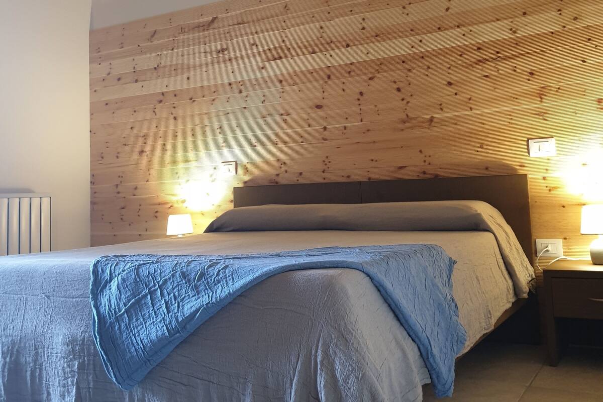 Makuuhuoneessa on puinen sängynpääty, harmaan ja sinisen värisellä peitteellä varustettu sänky, kaksi yöpöytävalaisinta ja yöpöytä.