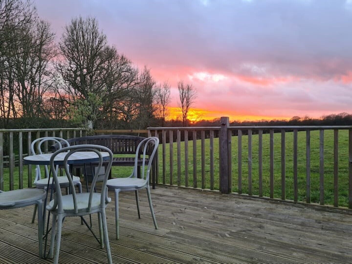 A wooden deck with metal chairs and a table, overlooking a grassy field with a wooden fence and trees.
