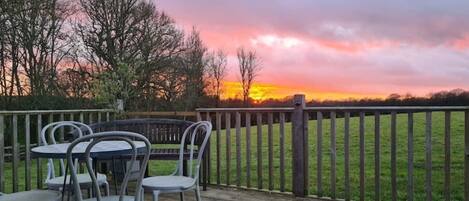 A wooden deck with metal chairs and a table, overlooking a grassy field with a wooden fence and trees.