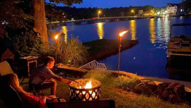 A lakeside evening scene with a fire pit, a dock, and a bridge in the background. - Lake Hopatcong cottage with dock; Permit #2023-6
(Hopatcong)