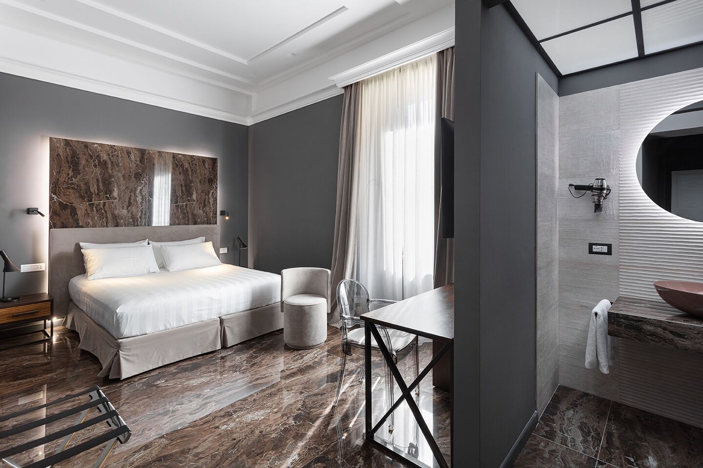 Habitación de hotel moderna con una cama grande, una mesa redonda, una silla, un tocador con espejo y un amplio ventanal con cortinas.