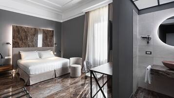 Habitació d'hotel moderna amb un llit gran, una taula rodona, una cadira, un tocador amb mirall i una finestra gran amb cortines.