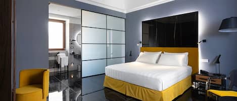 Quarto de hotel moderno com uma cama grande, uma poltrona amarela, uma escrivaninha com abajur e um espelho grande na parede.