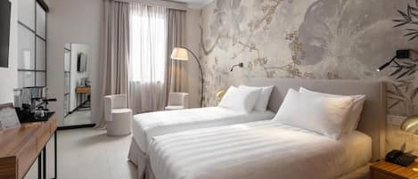 A hotel room with two beds, a desk, a chair, a lamp, and a wall with floral artwork.