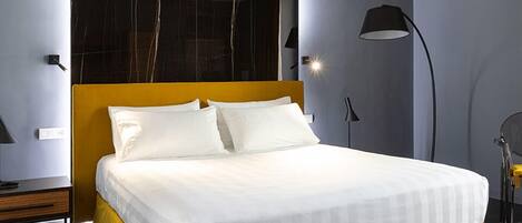 A modern hotel room with a large bed, a headboard with a mirror, a bedside table, and a floor lamp.