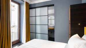 A hotel room with a bed, a window with curtains, a bathroom with a sink and mirror, and a dark wooden door. - Poerio 25 Boutique Stay (Naples)