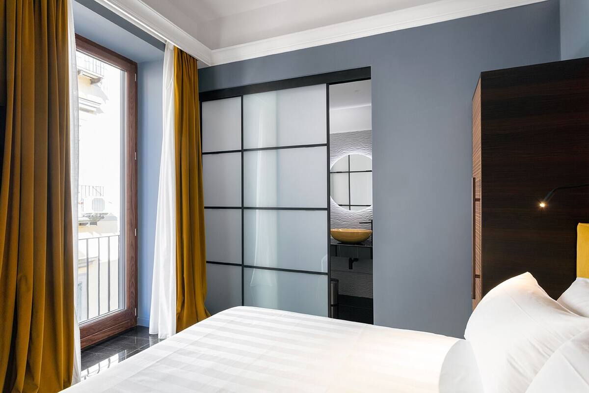 A hotel room with a bed, a window with curtains, a bathroom with a sink and mirror, and a dark wooden door.