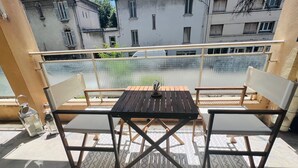 Ein Balkon mit einem Holztisch und zwei Stühlen, einem gemusterten Teppich und Blick auf Gebäude im Hintergrund.
