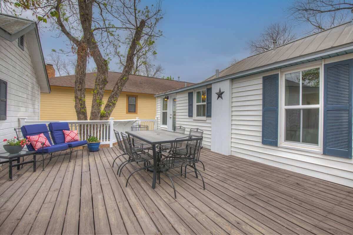 Une terrasse en bois avec une table et des chaises, un canapé bleu avec des coussins rouges, et une maison jaune en arrière-plan.