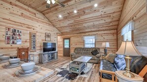 A cozy log cabin interior with a wooden ceiling, a television, a sofa, a coffee table, and a dining area with bowls and plates. - Cozy Cabin Firepit and Grill & 10 Mins to Main St (Fredericksburg)