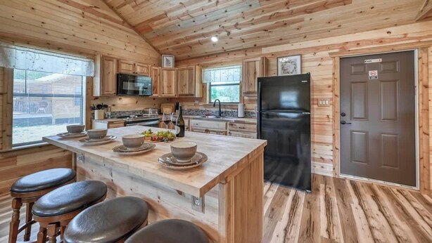 A wooden cabin-style kitchen with a central island, bar stools, a refrigerator, and a door with a sign. - Cozy Cabin Firepit and Grill & 10 Mins to Main St (Fredericksburg)