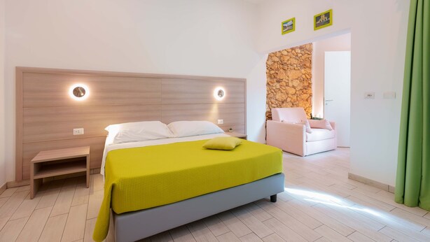 A modern bedroom with a bed, bedside table, and a bench. There is a stone wall and a door leading to another room. - Bardilio Luxury Rooms IUN P1813 (Cagliari)