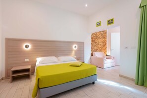 Un dormitorio moderno con una cama, una mesita de noche y un banco. Se observa una pared de piedra y una puerta que da a otra habitación.