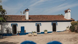 A white building with a tiled roof and blue trim, surrounded by a sandy area with potted plants.