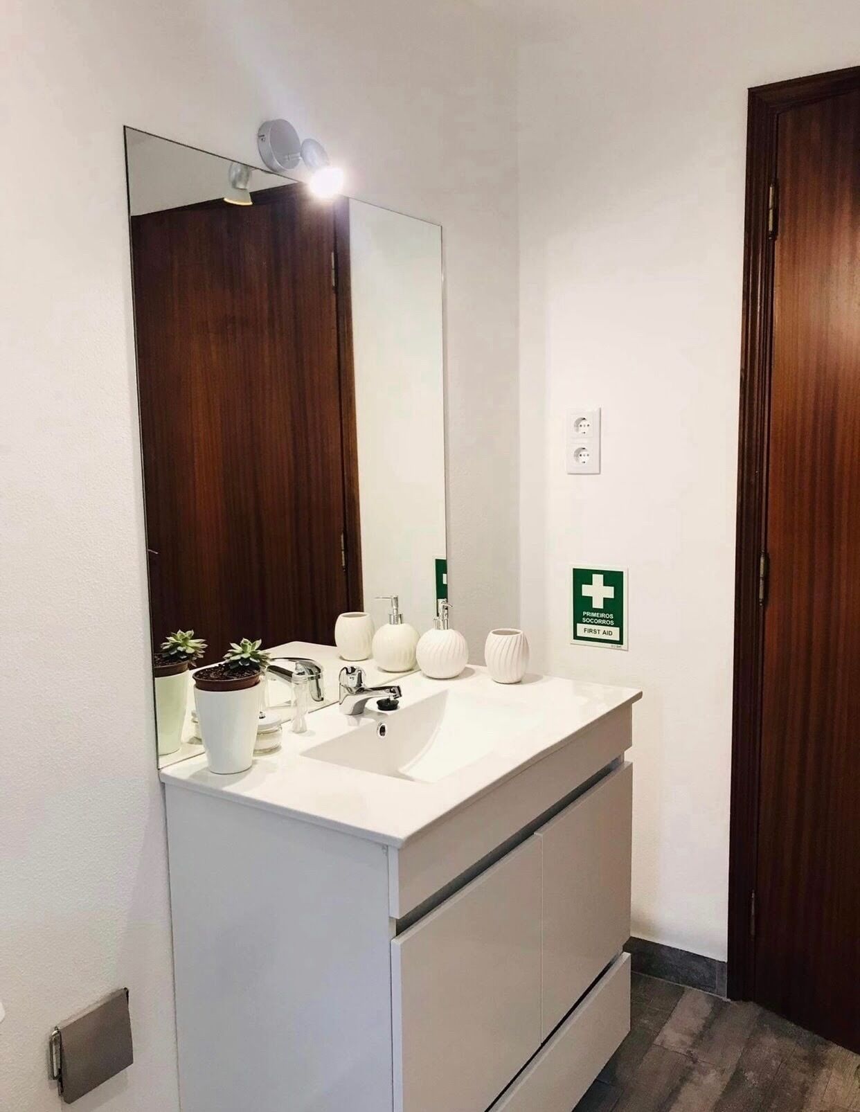 Een badkamer met een wit aanrecht, een grote spiegel, een wastafel en een groen bord met daarop het eerste hulpteken.