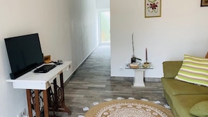 A living room with a flat-screen TV on a white table, a green sofa, a patterned rug, and a small side table with decorative items.