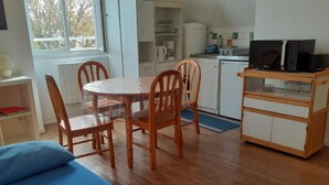 A compact room with a wooden dining table, two chairs, a bed with a blue cover, a kitchenette with a microwave, and a small cabinet. - Appartement de 38 m2 au Sein D'une Petite Résidence Avec Jardin (Pont-l'Abbé)