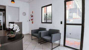 Un área de recepción con un mostrador, dos sillones grises, un reloj, un extintor y una ventana con vistas a un edificio.