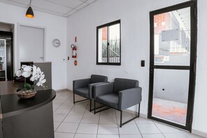 Un área de recepción con un mostrador, dos sillones grises, un reloj, un extintor y una ventana con vistas a un edificio.