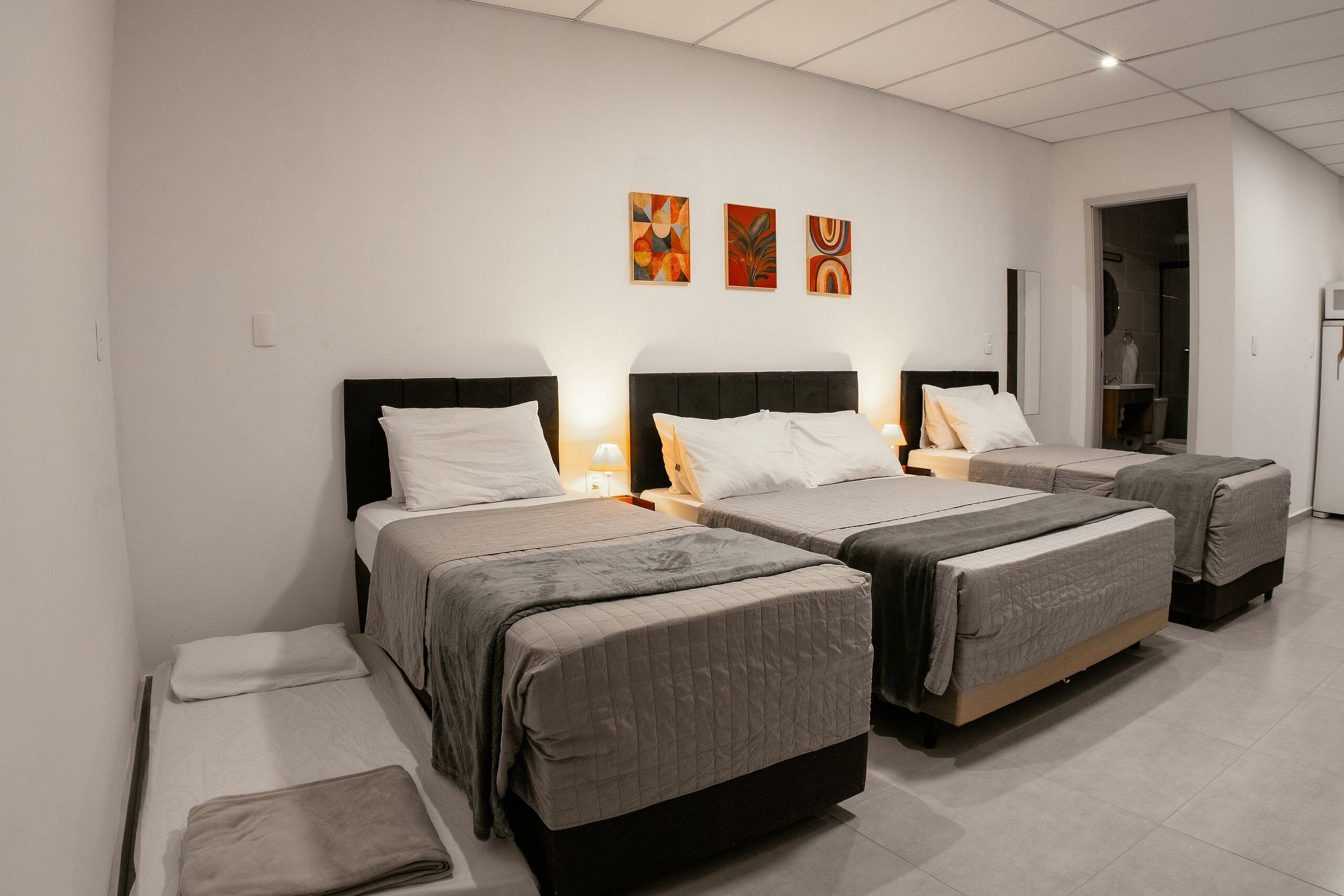 Quarto de hotel com três camas, banheiro e obras de arte na parede.