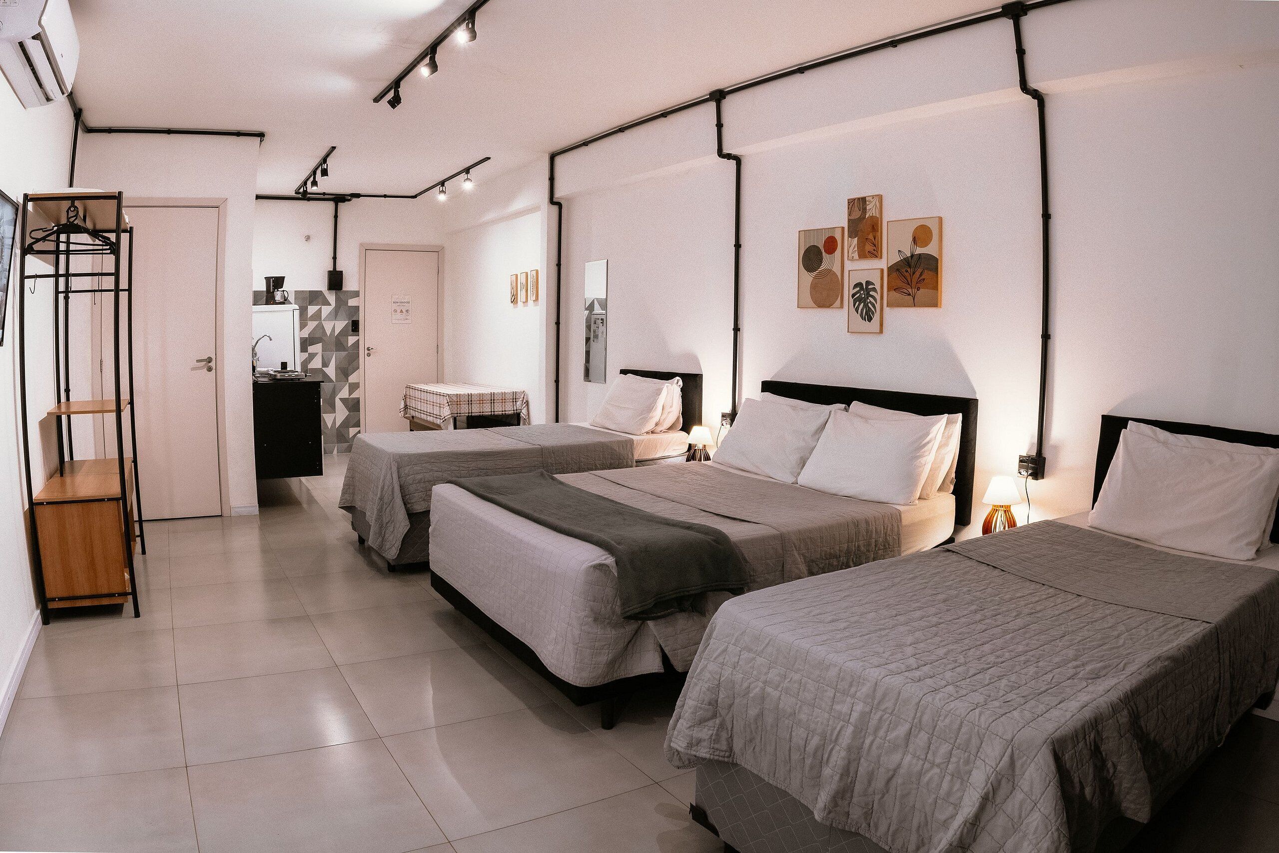 Ein Hotelzimmer mit drei Betten, einer kleinen Küchenzeile, einem Badezimmer und einem Kleiderständer.