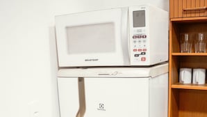 Een kleine keukenhoek met een witte koelkast, een magnetron erboven en een kastje met enkele spullen.