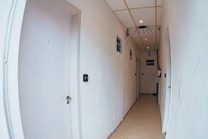 Un pasillo con paredes blancas, piso de baldosas y puertas. Se observan ventanas pequeñas y una lámpara en el techo.