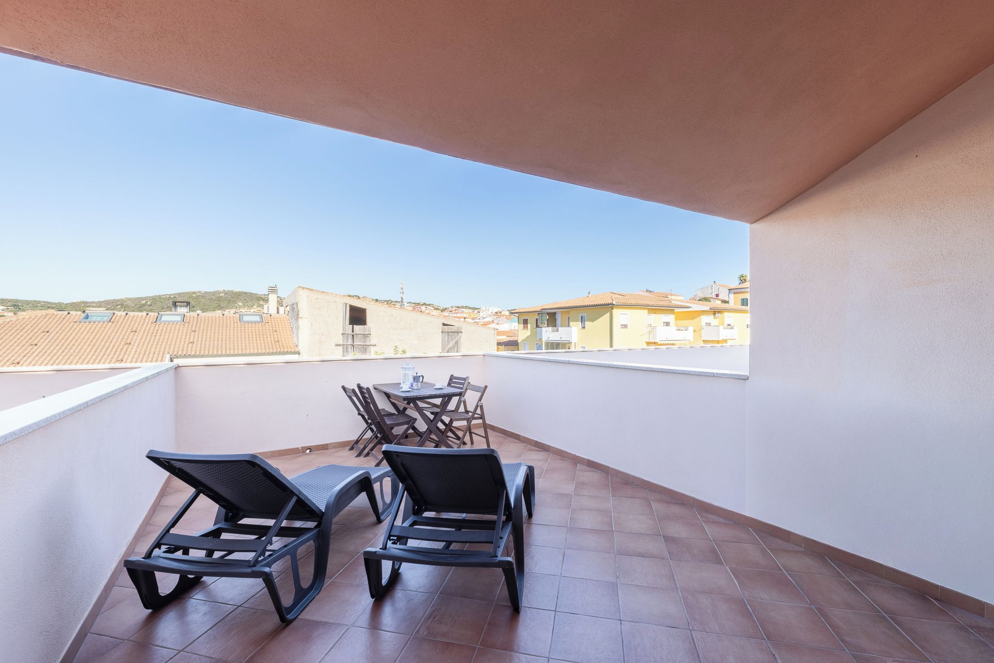 A balcony with two lounge chairs and a table, overlooking a residential area with tiled roofs.